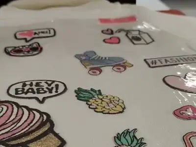 Blusa branca com apliques bordados cobertos pelo filme hotfix transparente, na mesa da prensa térmica